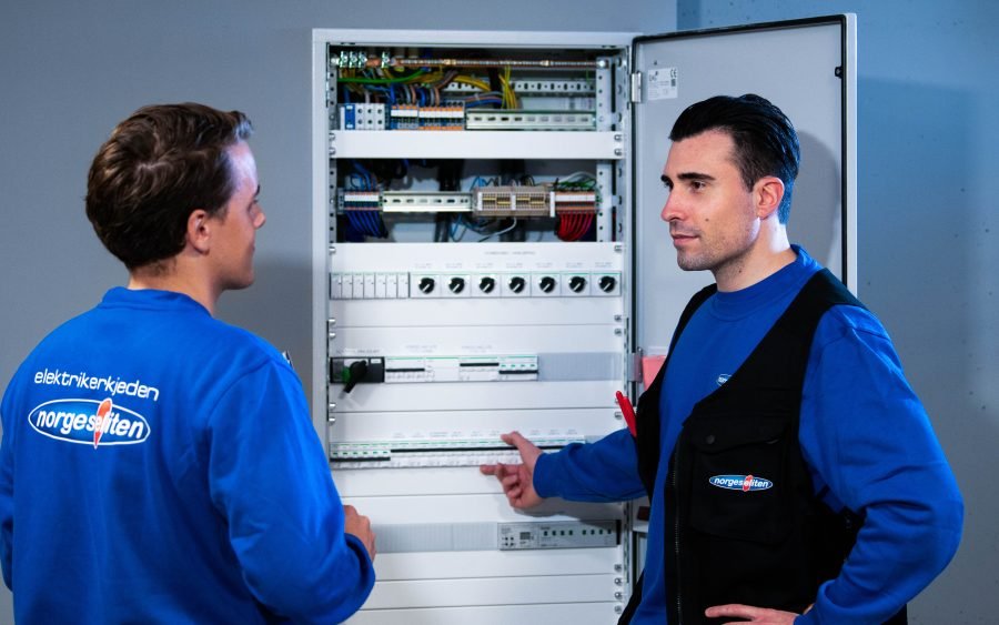 to unge mannlige elektrikere i blå arbeidsklær med Norgeseliten logo foran stort sikringsskap.